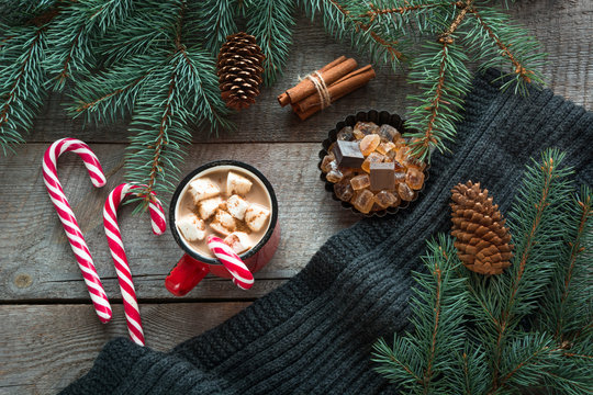 Mug hot coffee with milk, red candy cane on the wooden background with cone, branch of christmas tree and grey scarf. New Year. Holiday card. Rustic style. Top view and copy space.
