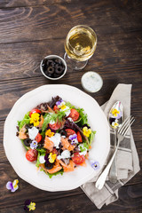 Fresh salad with smoked salmon, black olives, cherry tomatoes and edible flowers on wooden background. Top view.