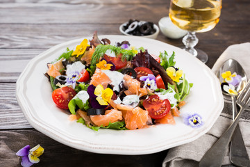 Fresh salad with smoked salmon, black olives, cherry tomatoes and edible flowers on wooden background.