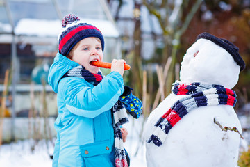 Funny kid boy in colorful clothes making a snowman, outdoors