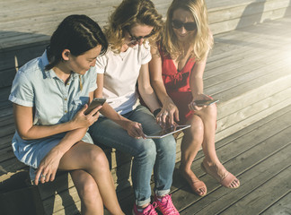 Three smiling girls sitting outside on wooden steps and using
 smartphones.One girl shows her friends pictures on screen tablet 
computer.Meeting friends.Young women using gadgets.