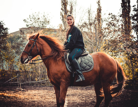 Young beautiful brunette girl rides a horse on a warm and sunny autumn day. Portrait of a pretty young woman on the horse, wearing tall boots and gloves.