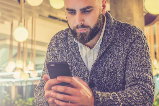 Young bearded businessman,dressed in gray cardigan,sitting at 
table in cafe and use smartphone.Man looks at screen of smartphone in 
his hands.Man using gadget.Guy browsing internet.