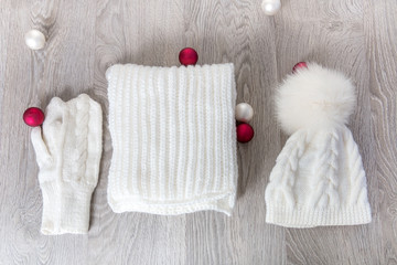 Knitted cold weather accessory near new year tree balls on the table. White hat, scarf ant gloves  grey wooden background. Top view. Christmas decoration.