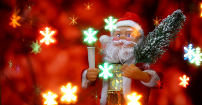 Santa Claus toy brings Christmas tree at glow red bokeh background and blurred lights snowflakes foreground. Big Copyspace concept New Year`s market banner, poster.
