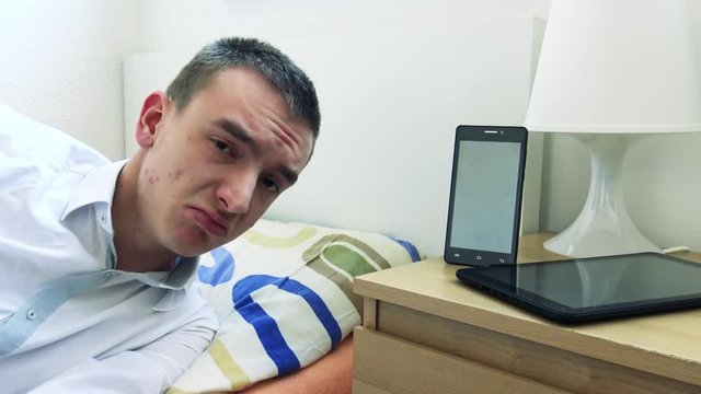 young man lying in bed watch to camera and man disagrees - smartphone and tablet