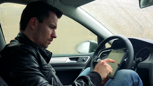 Young handsome serious man sits in car on place for driver and works on tablet 