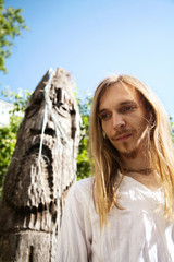 Slavic pagan young long-haired man next to a wooden pole idol sculprue