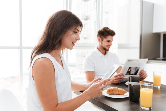 Woman having breakfast and using tablet while boyfriend reading newspaper