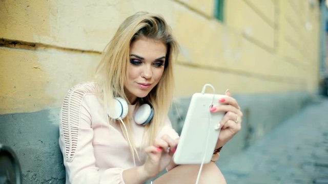 Blonde girl in elegant dress sitting in the alley and using tablet
