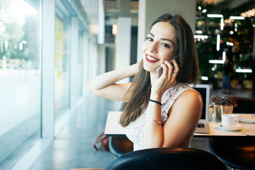 A charming female freelancer is talking on a phone while sitting in a cafe. Young attractive entrepreneur female is making a business call during the coffee break.