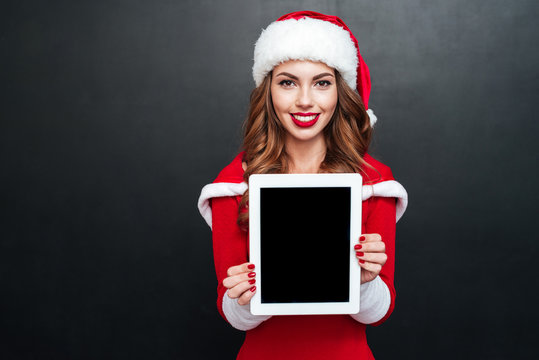 Cheerful woman in santa claus hat showing blank screen tablet