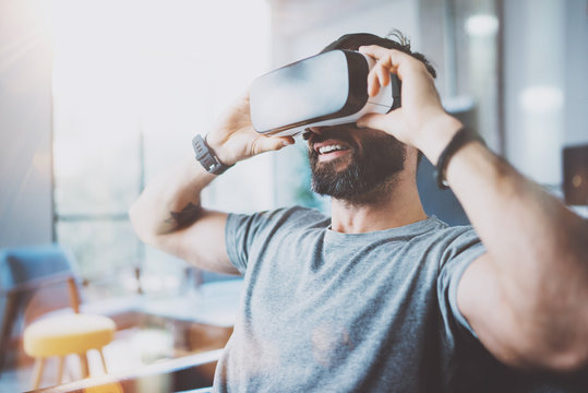 Bearded young man wearing virtual reality goggles in modern coworking studio. Smartphone using with VR headset in office. Horizontal, blurred.