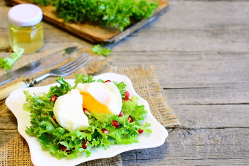 Poached egg, frisée lettuce leaves with pomegranate seeds and olive oil. Healthy salad on a plate, fork, knife on old wooden background with empty space for text. Tasty diet food. Closeup