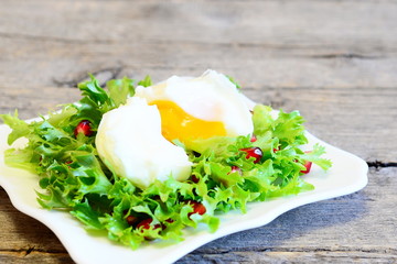 Poached egg, frisée lettuce leaves with pomegranate seeds and olive oil. Healthy salad on a plate isolated old wooden background. Tasty egg recipe. Closeup