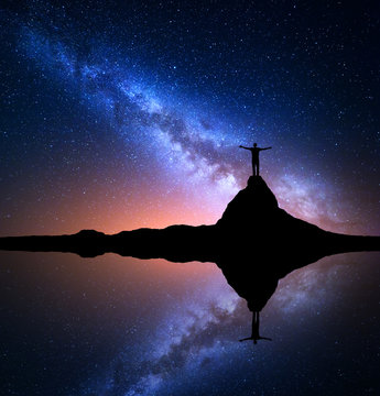 Milky Way. Night starry sky and silhouette of a standing happy man with raised up arms on the top of mountain near the lake with reflection in water. Milky Way and man on the rock. Galaxy, Universe