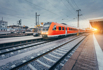 Obraz premium Modern high speed red commuter train at the railway station at colorful sunset. Railroad with vintage toning. Train at railway platform. Industrial landscape. Railway tourism. Vintage toning. Concept