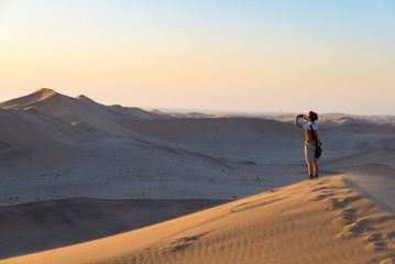 Tourist holding smart phone and taking photo at scenic sand dunes illuminated by sunset light in the Namib desert, Namib Naukluft National Park, Namibia. Adventure and exploration in Africa.