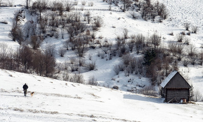 Hunter with a shotgun and a dog walking on snow-covered hill near the lonely wooden cabin. Winter hunting.