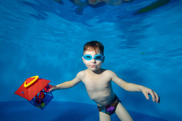Little boy swims underwater in the pool with a Christmas toy in hand and looking at me. Portrait. Shooting underwater. Landscape orientation - obrazy, fototapety, plakaty