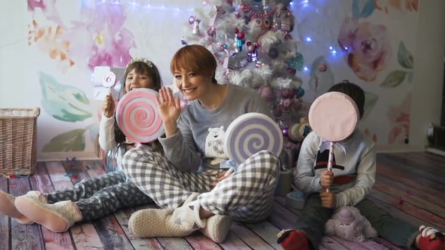Happy mother with her kids having fun with big toys candies before decorated christmas tree