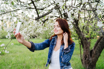 Young pretty smiling caucasian woman in jeans jacket and white dress taking selfie near blooming spring tree. Youth, freshness, beauty, happiness concept.