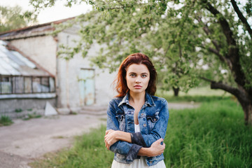 Young pretty serious caucasian woman in jeans jacket and white dress in a garden near the greenhouse. Youth, freshness, beauty concept.