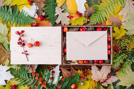 Autumn composition of colorful leaves and box filled with pines and small apples with blank craft envelope on wooden background. Top view, flat lay, copy space.