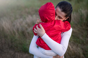 Portrait of beautiful young mother holding her baby toddler wearing red jacket. Happy family, mother and child play cuddling on field walk in nature outdoors.