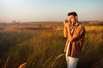Handsome young traveler man with vintage camera, outdoor take a picture of a meadow. Travel mood. Photography. Relaxation on a field and sunset background. Explore nature.
