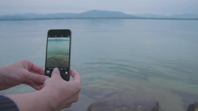 Young woman takes photographs of Lake Geneva using her smartphone camera. Close up on hands and phone.