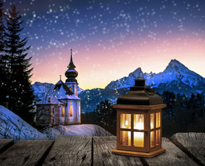 Laterne auf einem Holztisch vor verschneiter Winterlandschaft mit Kirche bei Nacht