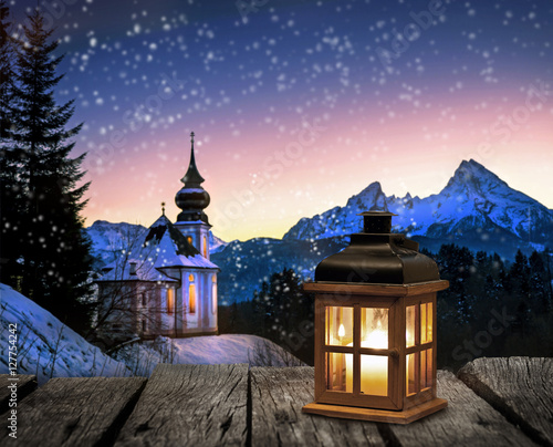 Fototapete Laterne Auf Einem Holztisch Vor Verschneiter Winterlandschaft  Mit Kirche Bei Nacht-Visions-AD