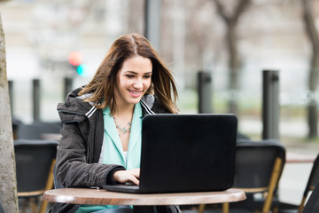 Busy young businesswoman is sitting in an outdoor caffe and using laptop. She is looking at camera and smiling.