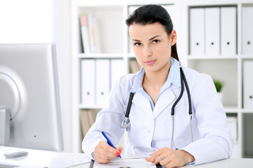 Young brunette female doctor sitting  with clipboard near window in  hospital and filling up medical history form.