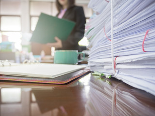 Pile of unfinished documents on office desk with businesswoman background