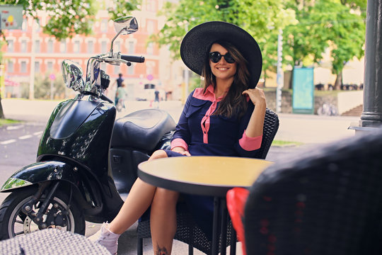 A woman in sunglasses and sunny hat sits at the table in a stree