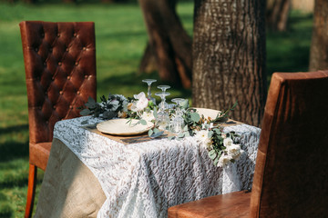 Dinner served for two outdoor, luxury lifestyle. Small table with two plates and silverware sets, decorated with green flowers, under tree with two cozy chairs