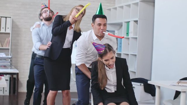Group of business people in party hats blowing horns and pushing chair with laughing woman while celebrating birthday in the office