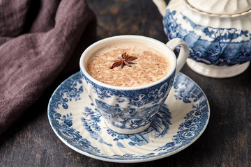 Masala tea chai latte traditional warm Indian sweet milk with spices, cinnamon stick, ginger herbs blend organic infusion healthy wellness beverage in porcelain cup on dark table background