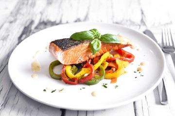Delicious salmon filet and red bell pepper vegetables on a white plate. Healthy meal, decorated with basil leaf.