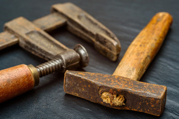 Old and well used handmade square hammer with short wooden handle and hand screw clamp on dark background.