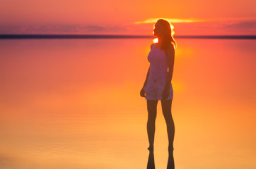 Beautiful female model stands in front of sun goes behind horizon at seaside. Salt lake Elton calm water during sunset reflects woman silhouette. Girl is alone