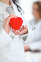 Female doctor with stethoscope holding heart.  Patients couple sitting in the background