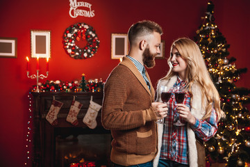 Attractive happy couple on Christmas day standing in front of the decorated tree smiling lovingly into each others eyes and holding glasses with red wine