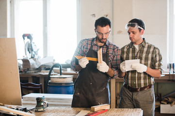 Happy male carpenter showing something to coworker at his notebook papers in workshop