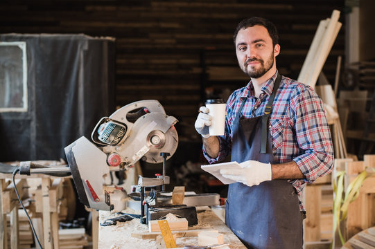 Carpenter taking a coffee break holding notebook in front of circular saw at his workshop