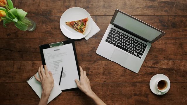 Man's arms typing on laptop checking papers writing Wooden table pizza slice aside cup of coffee Shot from above