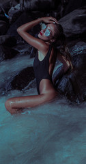 Beautiful glamorous woman in sexy black swimsuit and sunglasses standing and posing in lagoon sea water between shiny wet rocks