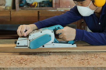 Carpenter using electric planer with wooden plank in carpentry workshop. He is wearing safety...
